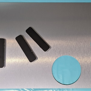 May include: A brushed silver metal plate with three black rectangular shapes and a circular cutout. The plate has a wavy edge on the right side.