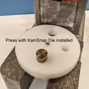 May include: A white press with a round white platform and a metal die. The platform has three holes and a small metal piece in the center. The text "Press with KamSnap Die installed" is visible below the platform.