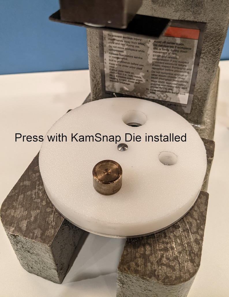 May include: A white press with a round white platform and a metal die. The platform has three holes and a small metal piece in the center. The text "Press with KamSnap Die installed" is visible below the platform.