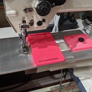 May include: A close-up of a sewing machine with a pink plastic attachment on the table. The attachment has two slots and a small knob.