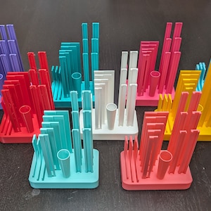 Può includere: Un set di nove portapenne geometrici stampati in 3D di colori vivaci. Ogni portapenne è di un colore diverso, tra cui viola, rosso, turchese, bianco e giallo. I portapenne sono disposti in uno schema sfalsato su una superficie di legno scuro.