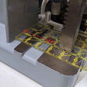May include: A yellow Omnigrid ruler with a grid pattern is positioned on a gray metal surface. The ruler is being pressed down by a metal bar.