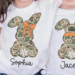 May include: Two white t-shirts with Easter bunny designs. One bunny has an orange bow, the other has an "Egg Hunter" hat. The bunnies are in camouflage colors. The names "Sophia" and "Jacob" are written below the bunnies.