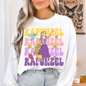 Pode incluir: Camisola branca de manga comprida com um gr&aacute;fico colorido de Rapunzel e a palavra "Rapunzel" em amarelo, rosa e roxo. O design inclui uma ilustra&ccedil;&atilde;o de desenho animado de Rapunzel, um p&aacute;ssaro azul e flores.