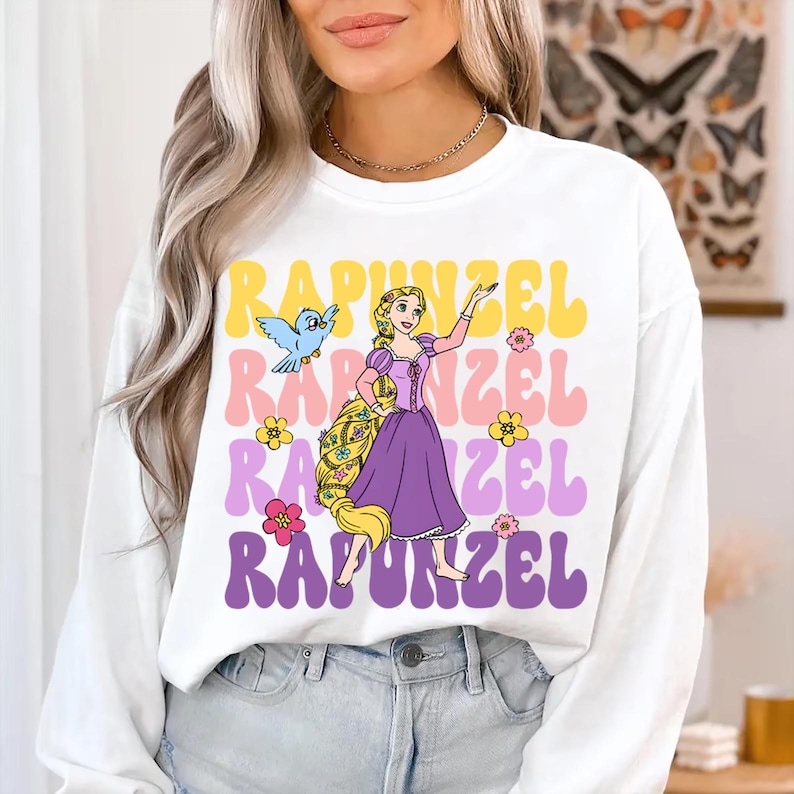 Pode incluir: Camisola branca de manga comprida com um gr&aacute;fico colorido de Rapunzel e a palavra "Rapunzel" em amarelo, rosa e roxo. O design inclui uma ilustra&ccedil;&atilde;o de desenho animado de Rapunzel, um p&aacute;ssaro azul e flores.