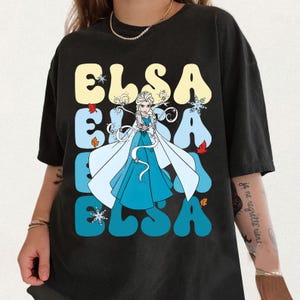 Pode incluir: T-shirt preto com o nome "ELSA" em uma fonte retr&ocirc; e um gr&aacute;fico de Elsa de Frozen. O design inclui flocos de neve e folhas de outono. A camiseta &eacute; de cor escura.