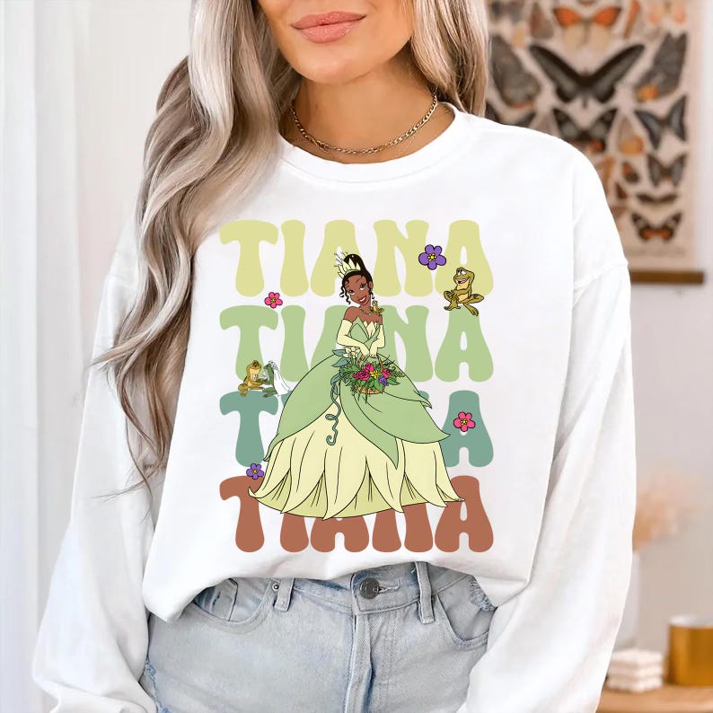 Pode incluir: Camisola branca com um gr&aacute;fico da Princesa Tiana em um vestido verde, com a palavra "Tiana" em letras retr&ocirc;. O design inclui sapos e flores. Uma blusa casual de manga comprida.