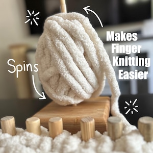 May include: A ball of thick, off-white yarn sits atop a small wooden board, with a wooden dowel in the center. The text "Spins" and "Makes Finger Knitting Easier" are visible. A row of wooden pegs is in the foreground.