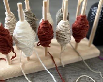 May include: A wooden yarn holder with multiple spools of yarn in various colors, including white, brown, and red. The yarn is hanging from the holder and is ready to be used for knitting or crocheting.