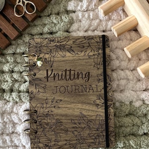 May include: A wooden knitting journal featuring floral engravings and the text "Knitting Journal". The journal has a spiral binding and a black elastic closure. A pair of scissors and a wooden knitting tool are also visible.