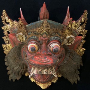 Puede incluir: Una máscara de madera roja con detalles dorados y blancos. La máscara tiene una expresión feroz con ojos grandes, dientes afilados y una corona detallada.