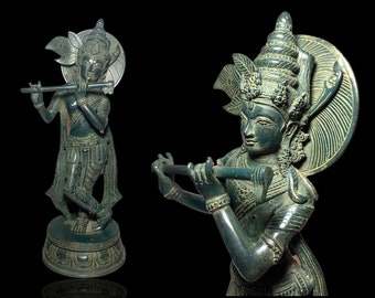 Op de afbeelding: Bronzen beeld van een godheid die een fluit bespeelt, met ingewikkelde details in kleding en hoofdtooi. Het beeld heeft een donkergroene patina en staat tegen een zwarte achtergrond. De figuur staat op een decoratieve basis.
