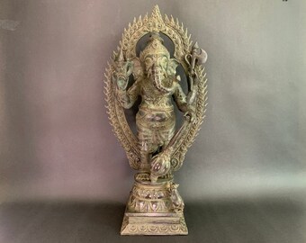 Op de afbeelding: Een bronzen beeld van de hindoe-god Ganesha, staand op een basis met een decoratieve halo. Het beeld is groen en heeft ingewikkelde details.