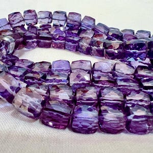 May include: A strand of faceted, square-cut amethyst beads. The beads are a deep purple color with lighter purple and white internal patterns. The beads are strung together, forming a curved shape. The background is a light, neutral color.