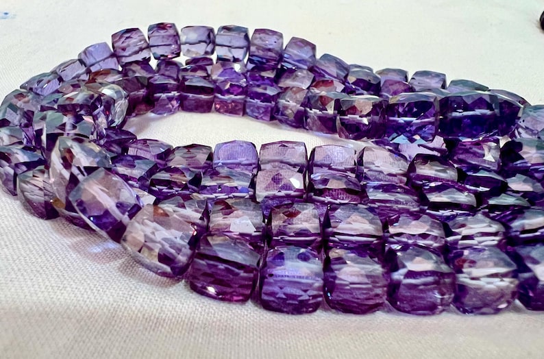 May include: A strand of faceted, square-cut amethyst beads. The beads are a deep purple color with lighter purple and white internal patterns. The beads are strung together, forming a curved shape. The background is a light, neutral color.