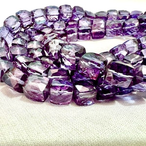 May include: A strand of faceted, purple amethyst beads. The beads are a mix of light and dark purple hues, with a geometric, rectangular shape. The beads are arranged in a circular pattern, resting on a white surface.