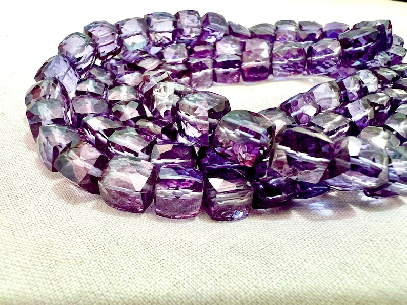 May include: A strand of faceted, purple amethyst beads. The beads are a mix of light and dark purple hues, with a geometric, rectangular shape. The beads are arranged in a circular pattern, resting on a white surface.