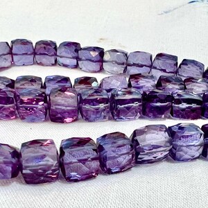 May include: Strands of faceted, purple gemstone beads. The beads are a mix of light and dark purple hues, with a square or rectangular shape. The beads are strung together, creating a necklace or bracelet.