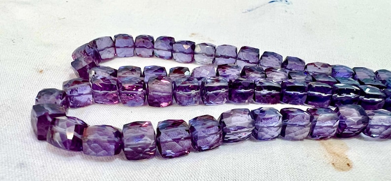 May include: Strands of faceted, purple gemstone beads. The beads are a mix of light and dark purple hues, with a square or rectangular shape. The beads are strung together, creating a necklace or bracelet.