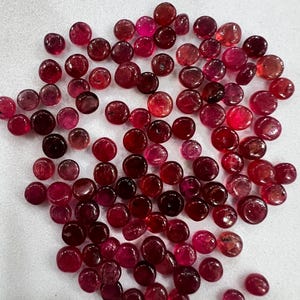 May include: A collection of numerous small, round, polished gemstones in various shades of deep red, crimson, and magenta. These smooth, circular stones are scattered across a plain, light-colored surface, displaying natural variations in color and translucency.