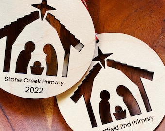 Könnte beinhalten: Zwei runde, weiße Holz-Weihnachtsornamente mit einer ausgeschnittenen Krippe. Das obere Ornament trägt die Aufschrift "Stone Creek Primary 2022" und das untere Ornament die Aufschrift "Westfield 2nd Primary 2022".