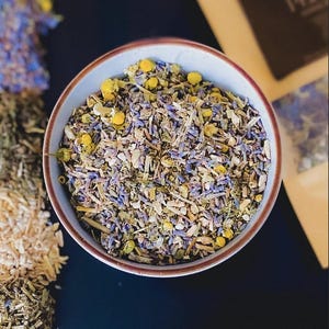 Puede incluir: Un cuenco de cer&aacute;mica lleno de una mezcla de hierbas y flores secas, incluyendo lavanda y manzanilla. El cuenco tiene un exterior azul claro con un borde marr&oacute;n. Otras hierbas y flores secas son visibles en el fondo.