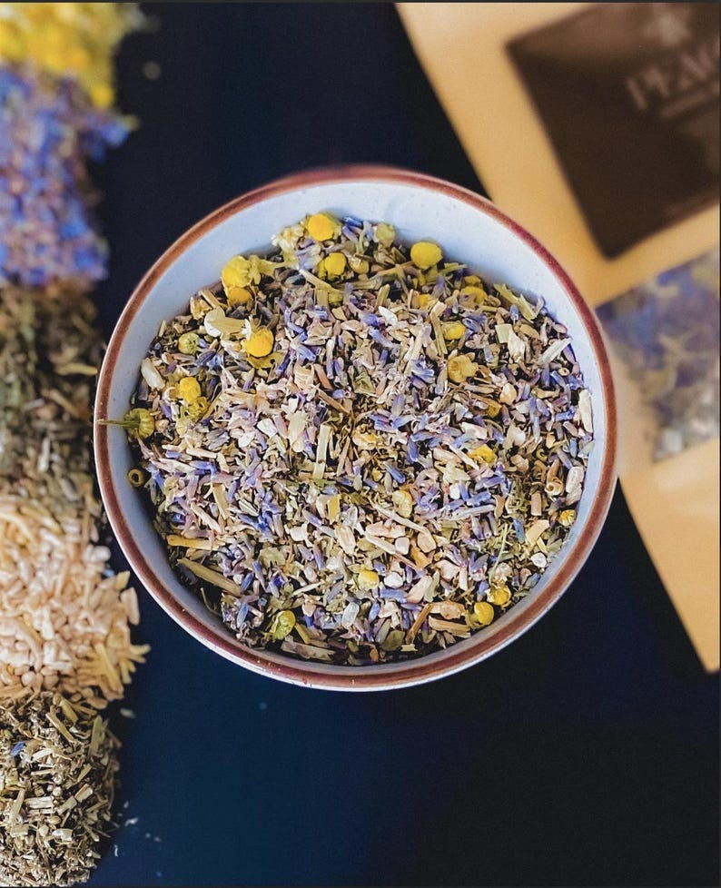 Puede incluir: Un cuenco de cer&aacute;mica lleno de una mezcla de hierbas y flores secas, incluyendo lavanda y manzanilla. El cuenco tiene un exterior azul claro con un borde marr&oacute;n. Otras hierbas y flores secas son visibles en el fondo.