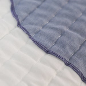 A close-up of two pieces of quilted fabric, one is light blue and the other is white. The light blue fabric has a scalloped edge and is sewn to the white fabric.