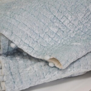 A light blue quilted throw blanket with a textured surface and pom pom trim.