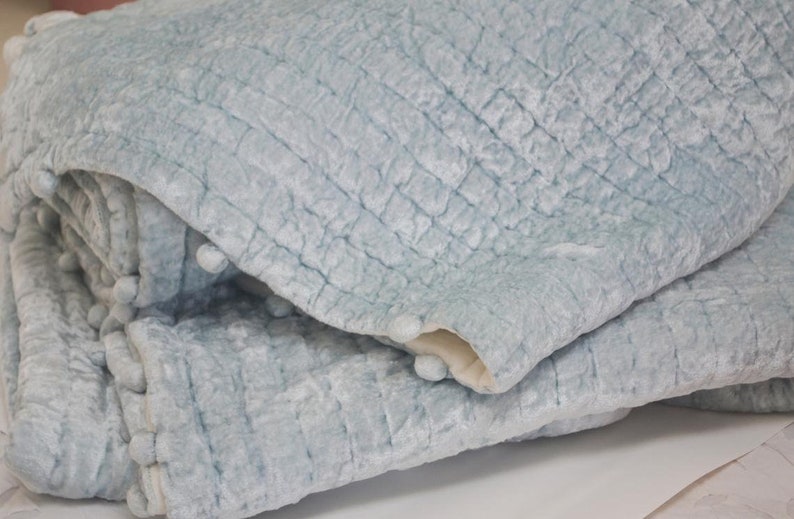 A light blue quilted throw blanket with a textured surface and pom pom trim.