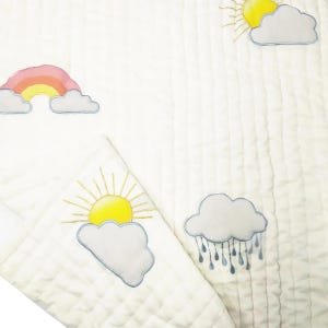 May include: A white quilted fabric with embroidered images of a rainbow, a sun, and a cloud with raindrops. The fabric has a subtle texture.