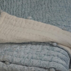 A blue and white quilted throw blanket with pom pom trim. The blanket is folded in half and the white side is facing up. The blanket is made of a soft, textured fabric.