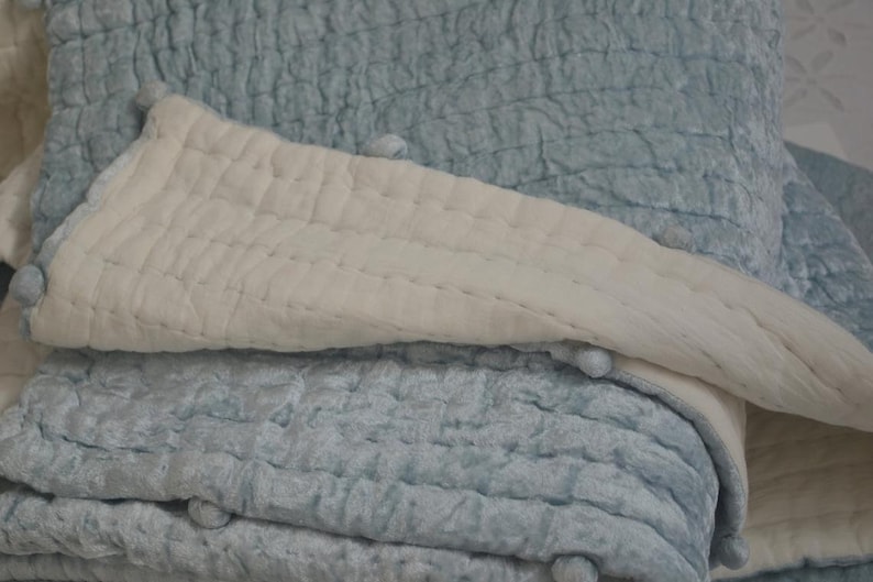 A blue and white quilted throw blanket with pom pom trim. The blanket is folded in half and the white side is facing up. The blanket is made of a soft, textured fabric.