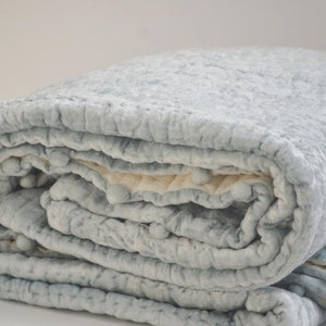 A neatly rolled icy blue velvet quilt showcasing its thick, inviting loft and ivory cotton reverse. This handmade quilt is pre-washed for extra softness, making it a favorite for a toddler blanket or a quilted baby blanket. Its durable construction ensures it works as a year-round throw quilt or a premium queen size quilt. Whether used as a decorative quilted throw or a personalized boy blanket, this velvet coverlet is a versatile and luxurious addition to any home.