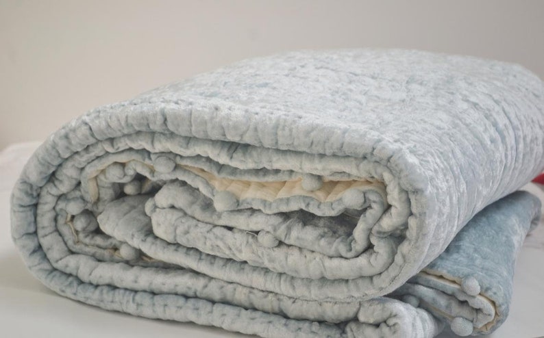 A neatly rolled icy blue velvet quilt showcasing its thick, inviting loft and ivory cotton reverse. This handmade quilt is pre-washed for extra softness, making it a favorite for a toddler blanket or a quilted baby blanket. Its durable construction ensures it works as a year-round throw quilt or a premium queen size quilt. Whether used as a decorative quilted throw or a personalized boy blanket, this velvet coverlet is a versatile and luxurious addition to any home.