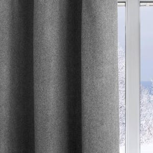 A lifestyle shot of a gray wool felt curtain hanging gracefully next to a window overlooking a snowy winter landscape. These thermal insulated drapes effectively block cold drafts, making them essential energy-saving curtains for a cozy home. The soft gray tone and heavy drape provide a minimalist farmhouse aesthetic, perfect for a nursery, kids room, or bedroom. This light-filtering wool curtain creates a warm and inviting atmosphere while functioning as a high-performance window treatment.