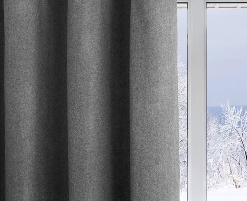 A lifestyle shot of a gray wool felt curtain hanging gracefully next to a window overlooking a snowy winter landscape. These thermal insulated drapes effectively block cold drafts, making them essential energy-saving curtains for a cozy home. The soft gray tone and heavy drape provide a minimalist farmhouse aesthetic, perfect for a nursery, kids room, or bedroom. This light-filtering wool curtain creates a warm and inviting atmosphere while functioning as a high-performance window treatment.