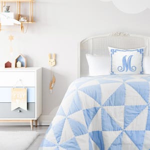 Può includere: Un piumino bianco e azzurro con un motivo geometrico. Il piumino è su un letto con una testiera bianca e un cuscino bianco con un cuscino monogrammato blu sopra. La stanza è decorata in uno stile chiaro e arioso con uno scaffale in legno e una decorazione da parete di coniglio di legno.