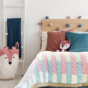 May include: A light blue, pink, and white striped quilt with a brown and white fox plush toy on top. The bed is made with a brown pillow and a teal blue pillow. A string of lights with paper lanterns hangs above the bed.