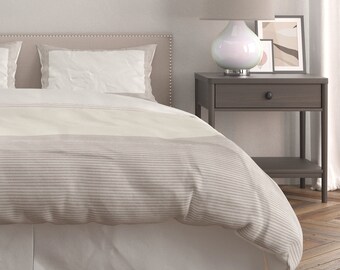 May include: A white and gray striped bedspread with a white bed skirt. The bed is made with two white pillows and a headboard with a light brown fabric. A nightstand with a drawer and a shelf is next to the bed. A white lamp with a round base is on the nightstand.