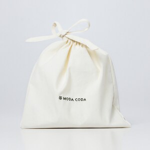 以下が含まれることがあります： 緑色の「MODA CODA」の文字が印刷された、白い綿の巾着袋です。
