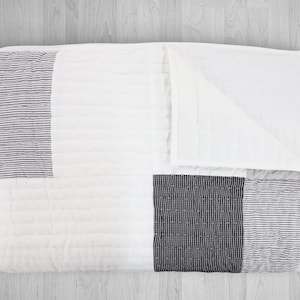 Peut inclure: Une couette blanche avec un motif de patchwork composé de rayures noires et blanches. La couette est pliée en deux et a une surface texturée.
