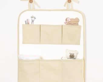 May include: A beige fabric hanging organizer with six pockets. The organizer has two wooden dowels at the top and is hung on a wall. The pockets are filled with various items, including a stuffed animal, a toy, and a towel.