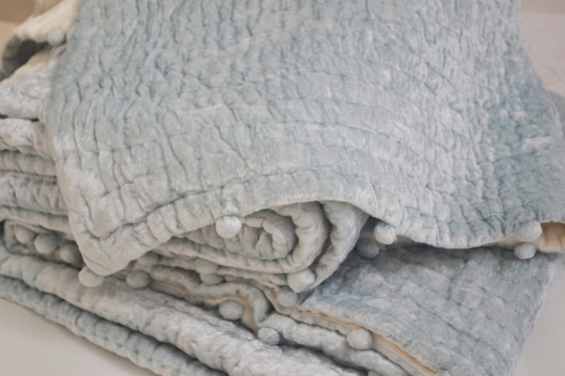 This close-up highlights the plush texture of the blue-gray velvet pick stitch quilt. The medium-weight handmade blanket is filled with soft cotton batting, functioning as a warm bed quilt or lap quilt. Available in custom sizes from crib quilt to king size quilt, it serves as a high-quality baby shower gift or a stylish twin quilt for students. The tonal depth and pom pom quilt trim make it a standout piece for a bohemian or modern bedding set.