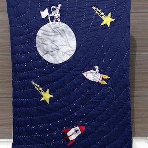 以下が含まれることがあります： 月面に宇宙飛行士、ロケット、流れ星が描かれた青いキルトの毛布。毛布は白いステッチで飾られています。