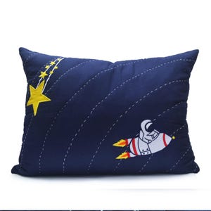 May include: A blue pillow with a white rocket ship and a yellow star with a trail of smaller yellow stars. The pillow is quilted with a white thread.