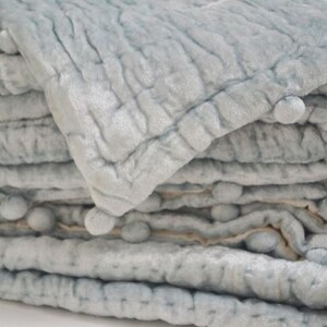 A luxurious icy blue velvet quilt featuring a hand-quilted pick stitch pattern and playful pom pom edges. This handmade velvet coverlet is backed with soft ivory cotton, making it a perfect bedspread or comforter. The rich quilting fabric offers a modern quilt look with the cozy feel of a vintage quilt. Ideal as a dorm bedding set or a sophisticated throw blanket, this breathable quilted blanket adds a touch of elegant home decor to any bedroom.
