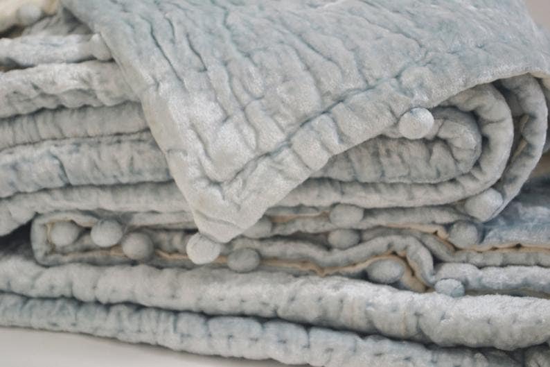 A luxurious icy blue velvet quilt featuring a hand-quilted pick stitch pattern and playful pom pom edges. This handmade velvet coverlet is backed with soft ivory cotton, making it a perfect bedspread or comforter. The rich quilting fabric offers a modern quilt look with the cozy feel of a vintage quilt. Ideal as a dorm bedding set or a sophisticated throw blanket, this breathable quilted blanket adds a touch of elegant home decor to any bedroom.