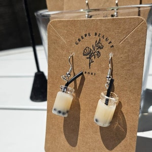 May include: A pair of dangle earrings featuring miniature boba tea drinks. Each earring is a clear jar filled with a creamy liquid and dark boba pearls, with a black straw. The earrings are displayed on a brown card with the text "CREPE BEAUTY".