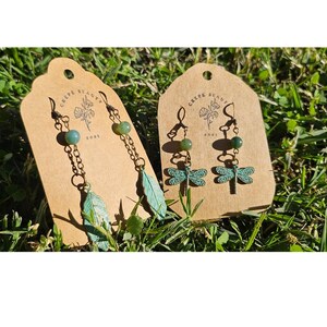 May include: Two pairs of handcrafted earrings displayed on brown tags. One pair features a teal feather charm, the other a dragonfly. Both have green beads and are suspended from delicate metal chains. The tags have the text "Crepe Beauty" and a floral design.